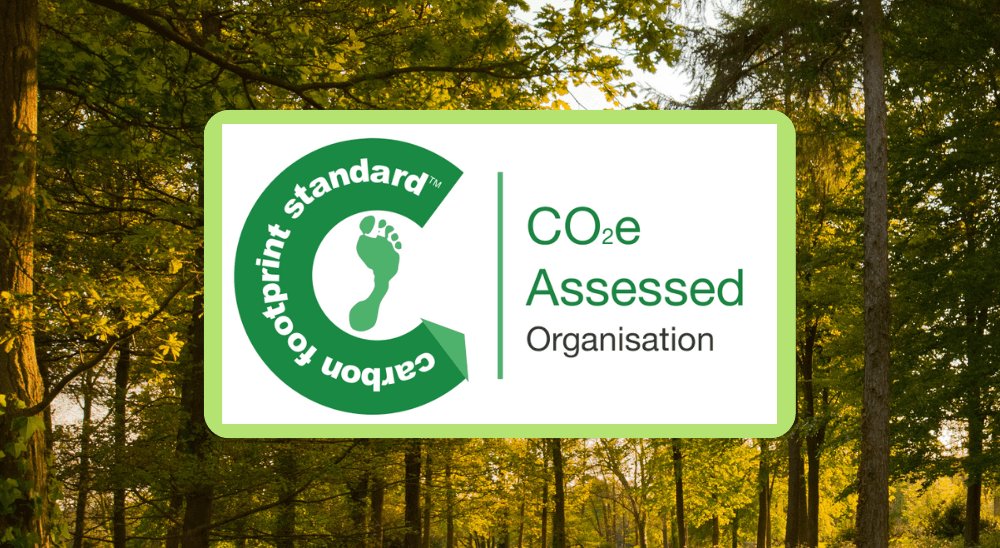 The image shows the Carbon Footprint Standard logo, featuring a green footprint inside the letter "C," with the text "CO2e Assessed Organisation" next to it. The background is a serene, sunlit forest with green trees.