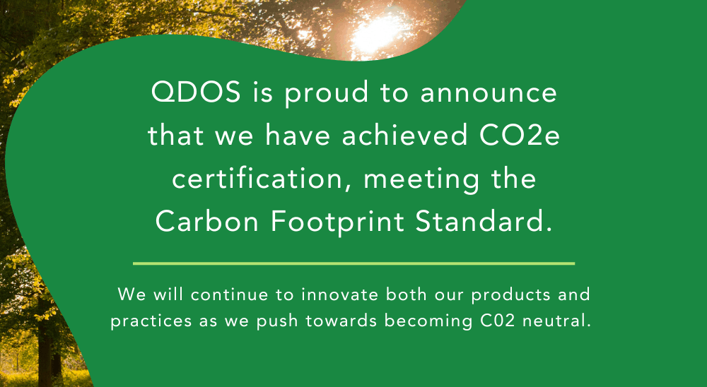 The image contains a green background with white text stating: "QDOS is proud to announce that we have achieved CO2e certification, meeting the Carbon Footprint Standard." Below, it adds, "We will continue to innovate both our products and practices as we push towards becoming CO2 neutral." The background features a forest scene with sunlight breaking through the trees.